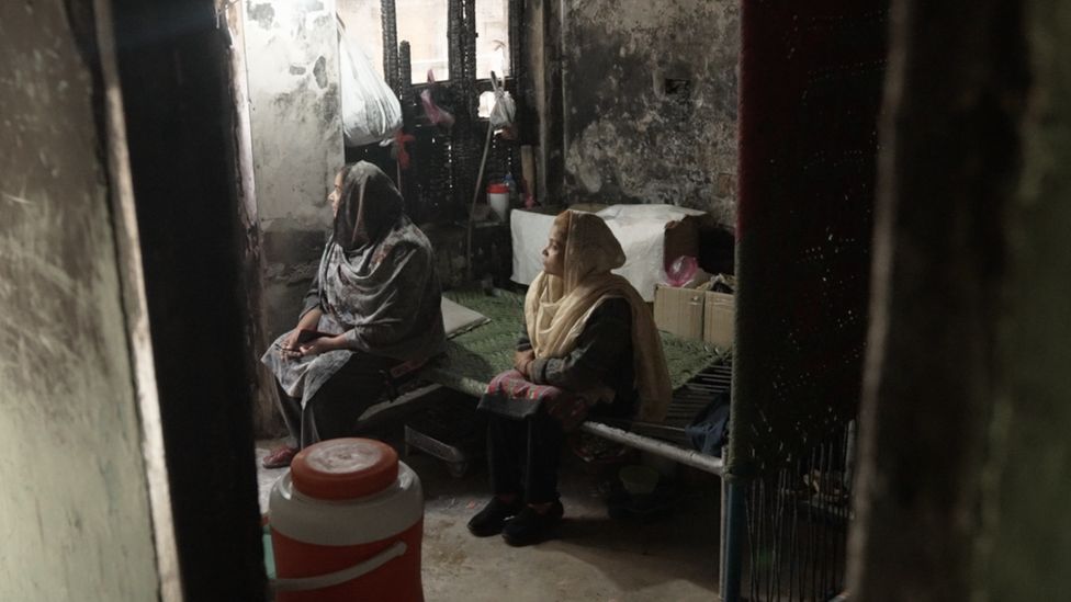 Sima sits on a bed in her house next to another women, both looking out of a window