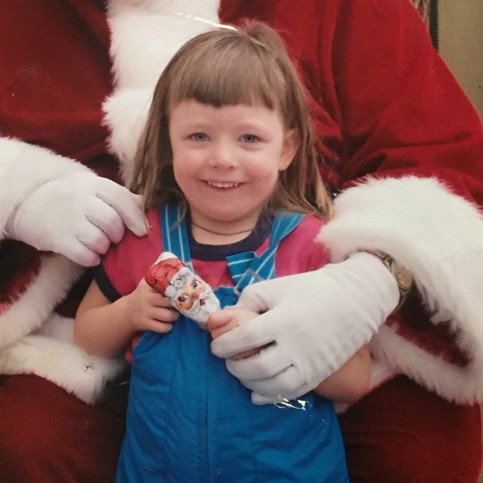 A young Ashley at around 7-years-old standing in front of a Santa Claus