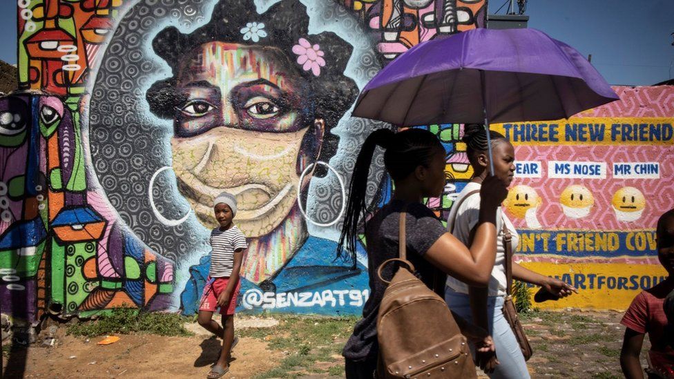 People walking past a mural of a woman with a mask. She has hoops on and a bantu hair style. It is a graffiti mural.
