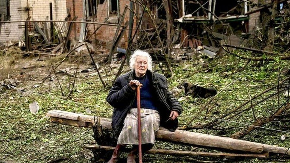 A woman sits on a fallen log in front of two houses that have been demolished