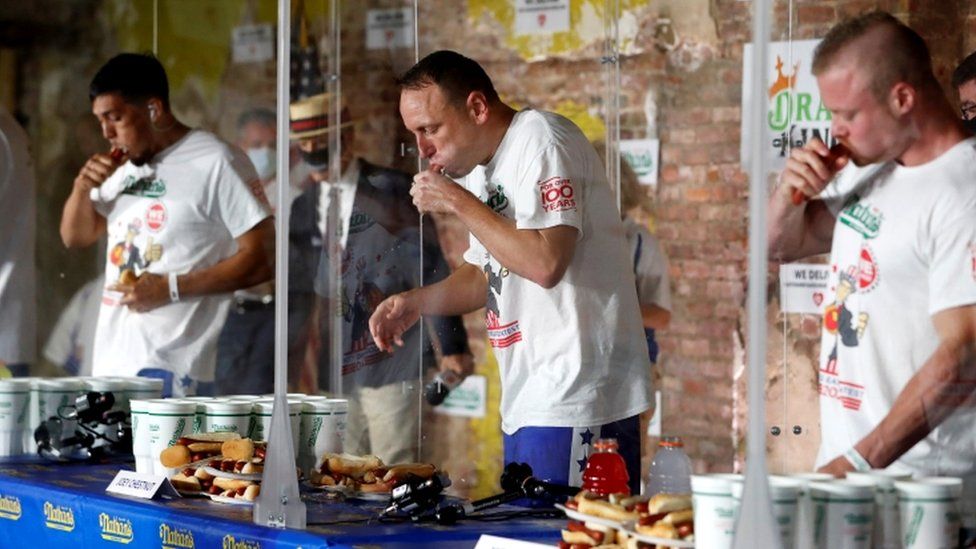 Джоуи Честнат (в центре) соревнуется с участниками, разделенными плексигласом, в Международном конкурсе по поеданию хот-догов Nathan's Famous Four of July
