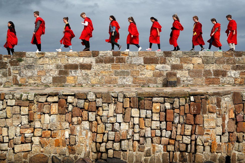 A row of students in red gowns walk along the harbour wall.