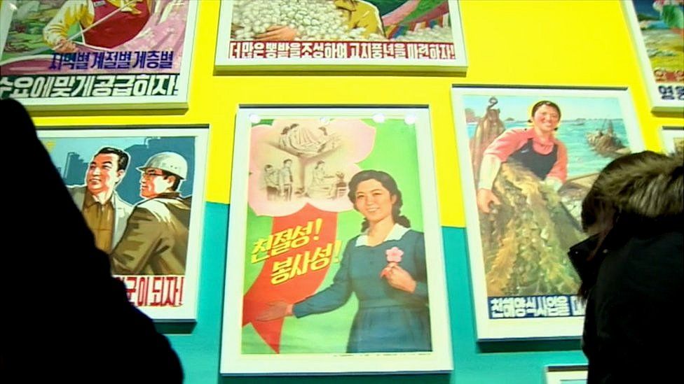Visitors are staring at North Korean posters. On the centre poster with fluorescent green coloured background, a young woman is emphasising the importance of kindness and servitude.
