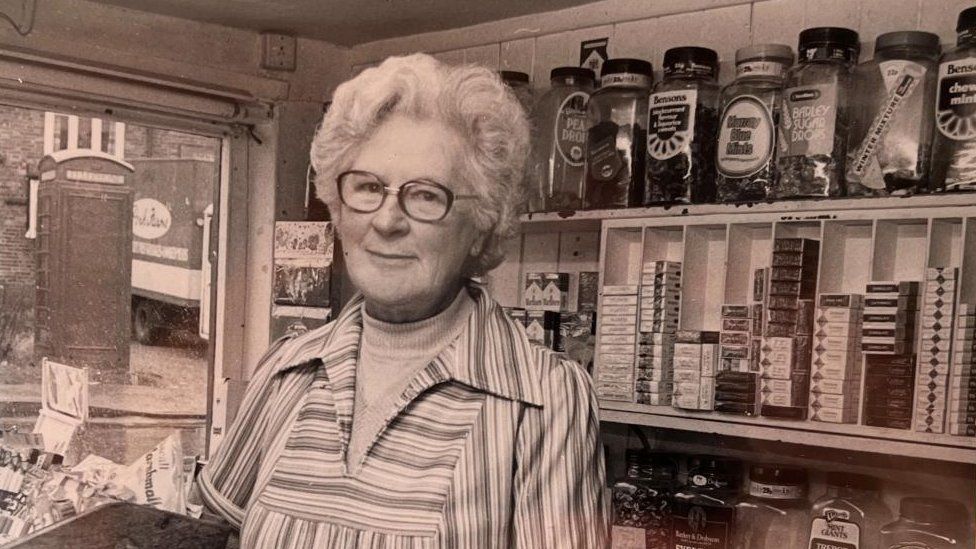 Black and white photo of elderly woman with glasses standing behind the counter of a shop with jars of sweets behind her