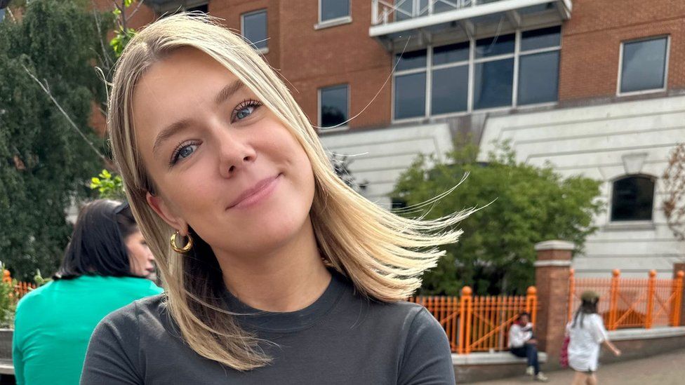 Kali Thompson. Kali is a 25-year-old white woman with shoulder length blonde hair and blue eyes. She wears a stone grey T-shirt and gold hoop earrings. She's pictured outside in front of a building with children playing nearby