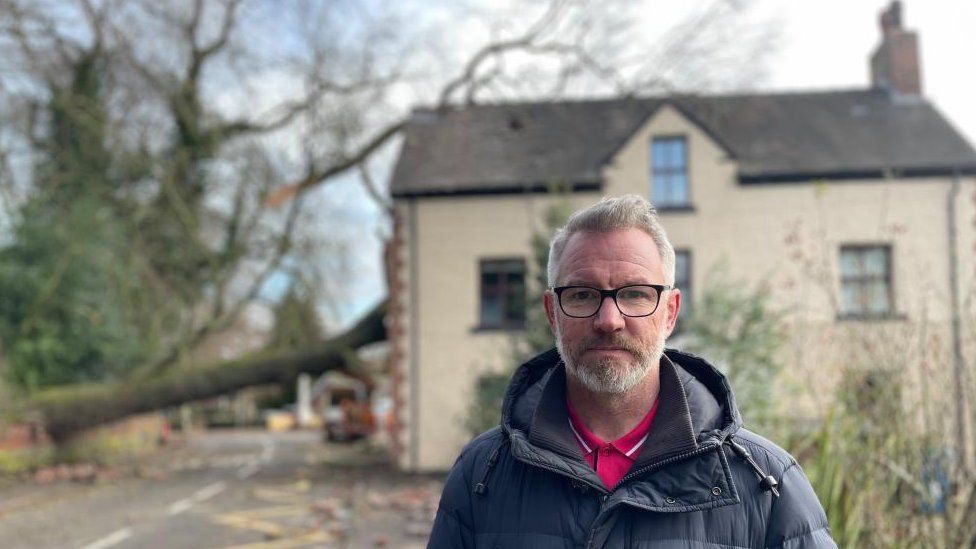 A man standing in front of his home which a tree crashed into