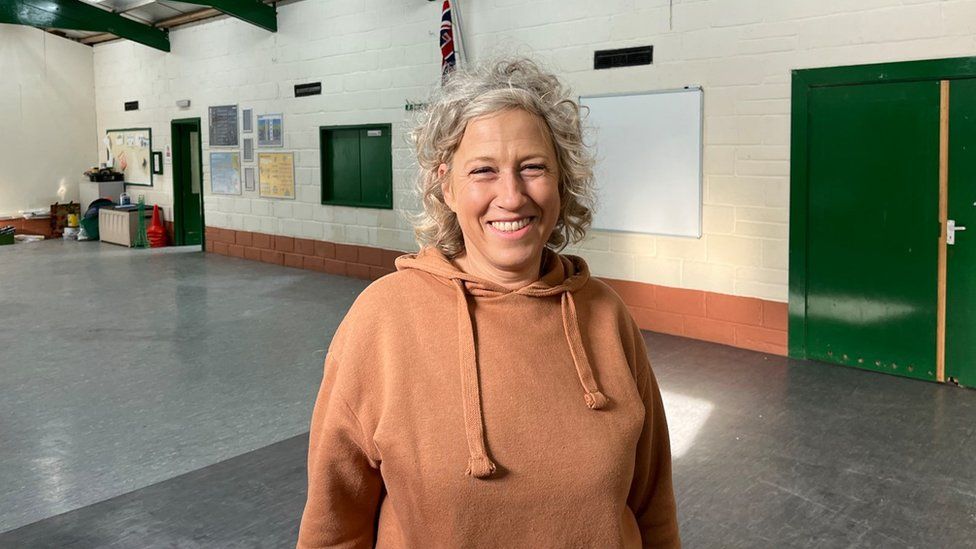 Fitness instructor Claire Webber has grey hair and stands inside the redecorated scout hut smiling at the camera wearing a brown hoodie