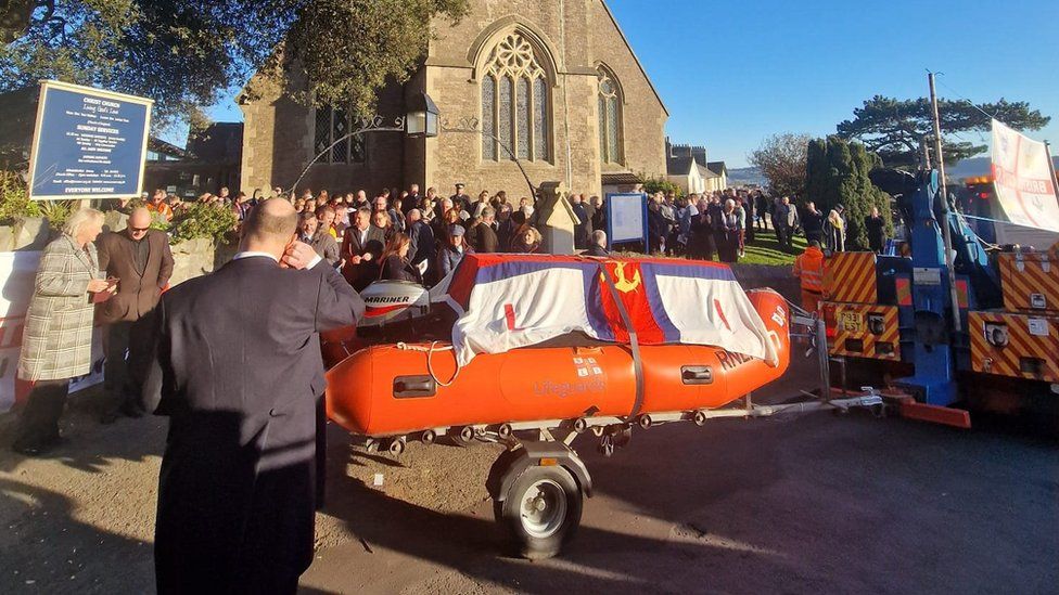 The coffin on a lifeboat outside a church with mourners nearby in Weston-super-Mare