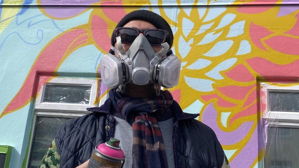 A street artist in a fume mask holding a can of pink spray paint in front of a wall spray painted with bright flowers