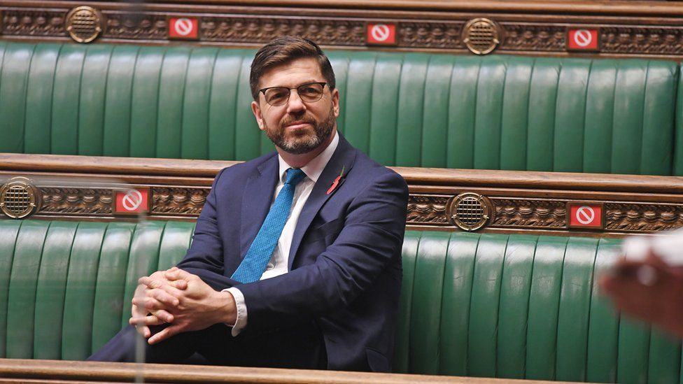Conservative MP Stephen Crabb sitting on the green benches in the House of Commons., hands clasped around one knee smiling