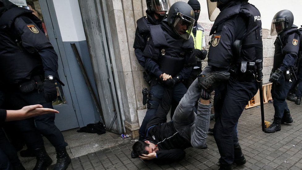 A man, lying on the ground, clutches his head in pain as he is dragged through the street by his legs by two police officers in full riot gear