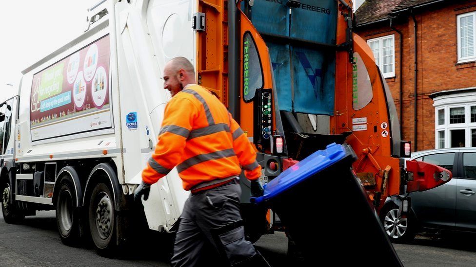 A refuse collector wheels a bin towards a bin lorry