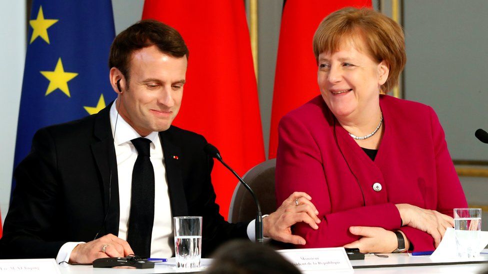 Mr Macron, smiling to himself, places a hand on Mrs Merkel's arm while she laughs at something as the pair sit at a table during a joint news conference