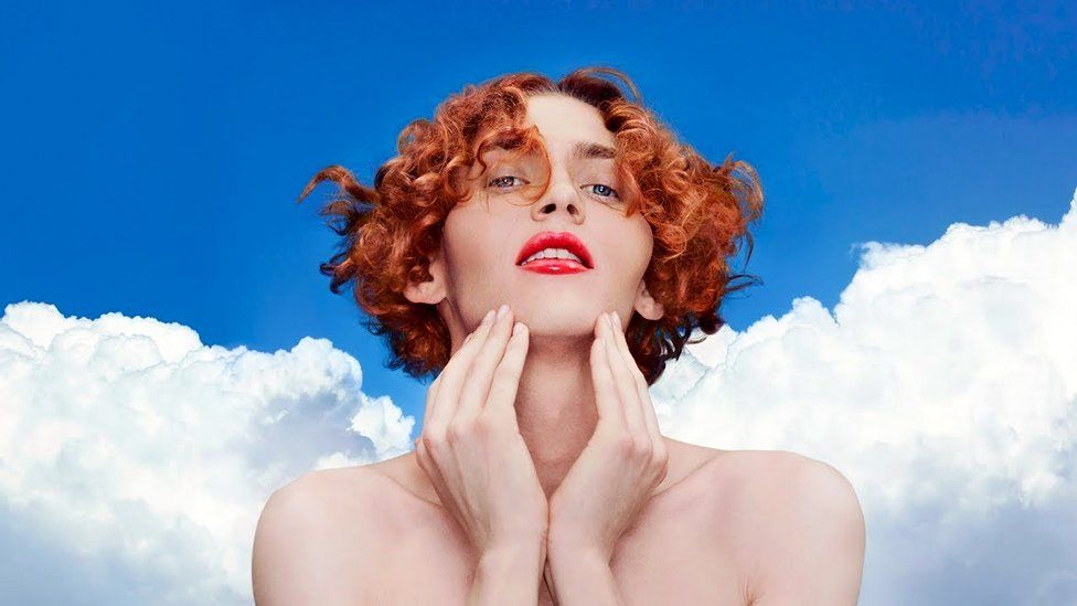 A head and shoulders shot of the producer and singer, Sophie, with her signature short red curly hair, holding her hands on her neck. The background features a blue cloudy sky.