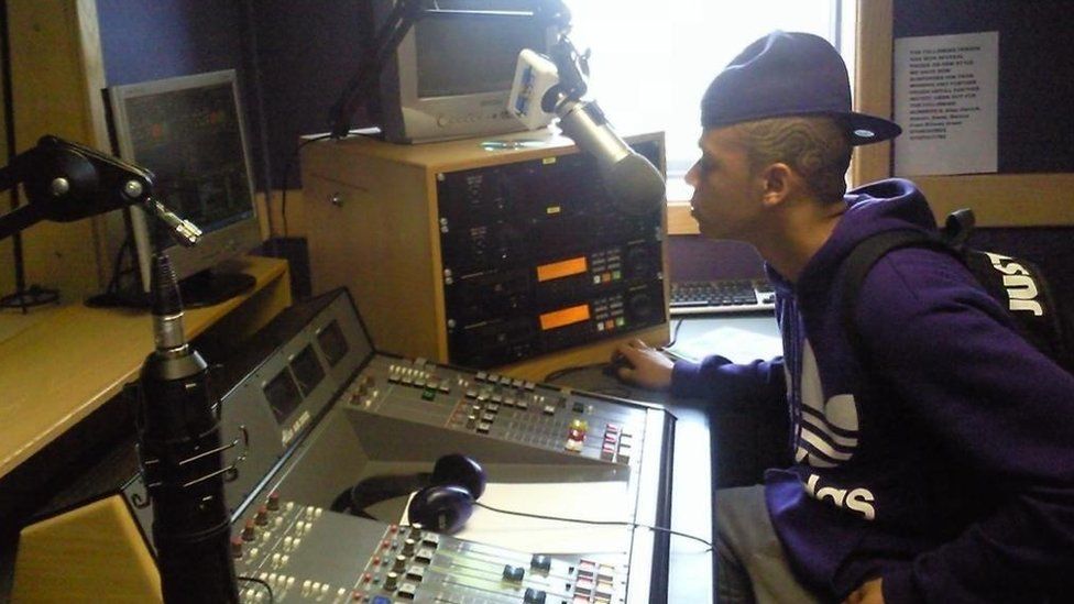 DJ Day Day when he was aged 14 sitting in a radio studio. He is sat on a chair with his right hand on a computer mouse and a radio mic in front of his mouth. In front of him is a radio desk with lots of computing equipment for mixing music and also a computer monitor. He is wearing blue cap backwards on the top of his head and is wearing a blue hooded top with the Adidas log on the front. He is also wearing a black Nike rucksack on his back.