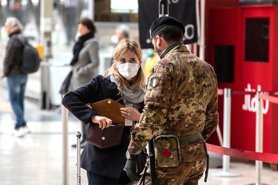 A woman wearing a face mask speaks to a soldier