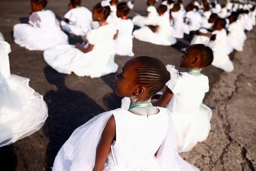 Young girls sitting on the ground in rows wearing white dresses.
