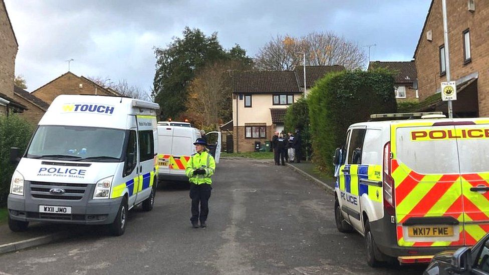 Image of Wedmore Close in Kingswood. Police vans can be seen either side of the road, and a female officer is standing next to a van. Houses can be seen either side of the road and in the distance.