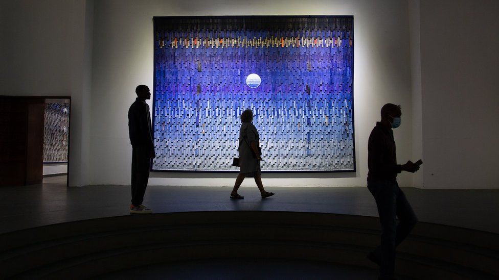 Viewers walk past work by Malian artist Abdoulaye Konaté at the Ancien Palais de Justice on May 21, 2022 as part of the Dakar Biennale. The artwork features varying shades of blue and contains what looks like the moon.