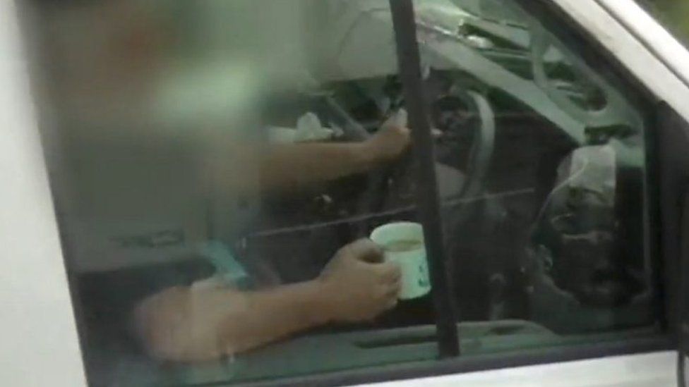 Motorist sat in driver's seat clutching cup of tea with his right hand and steering wheel with his left