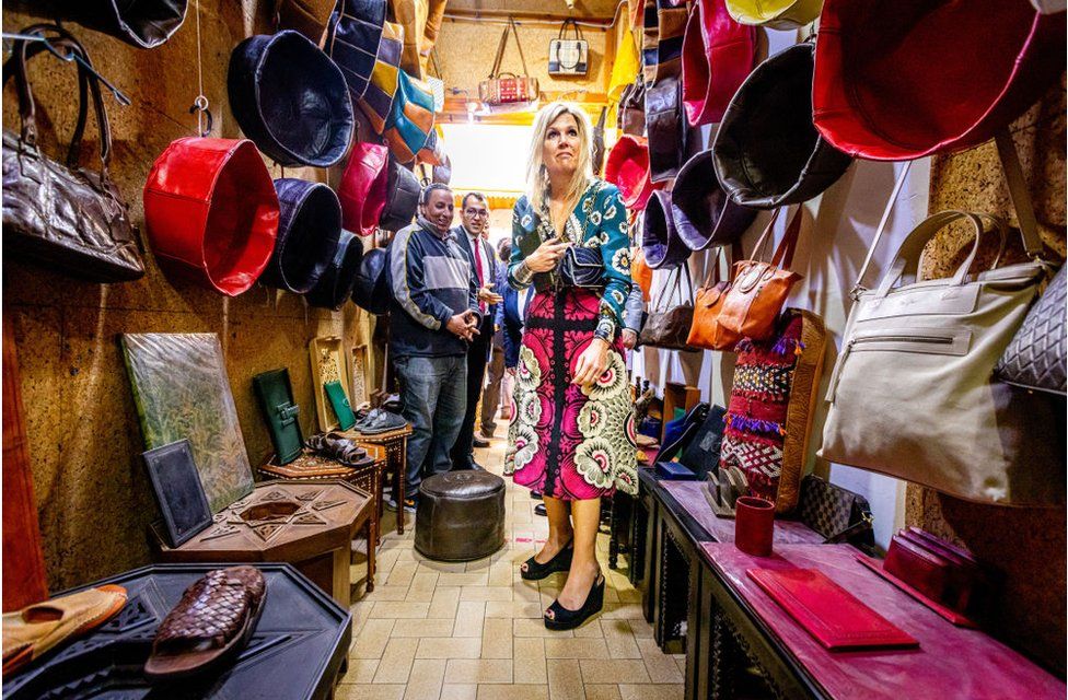 Queen Maxima in a hat shop looking at multicoloured hats which are hanging from the walls.
