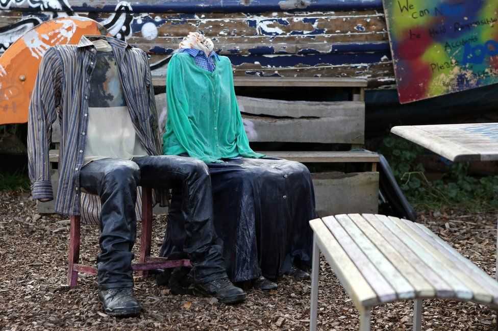 Two chairs are dressed in clothing, one in shirt and trousers and the other in a skirt and a cardigan