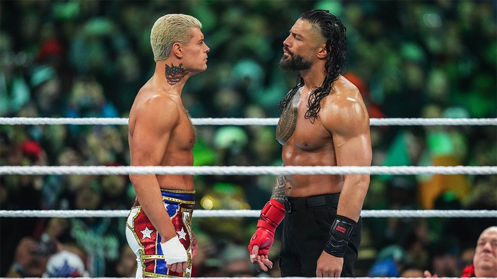 Cody Rhodes and Roman Reigns face off. Cody is a 38-year-old white man with short, bleached blonde hair and Roman is a 38-year-old Samoan man with long, curly brown hair. Both wrestlers are shirtless exposing their muscly chests - Roman's chest is covered in a large tattoo while Cody has one on the right side of his neck. Cody's shorts are the red, white and blue of the US flag complete with stars while Roman's are black.