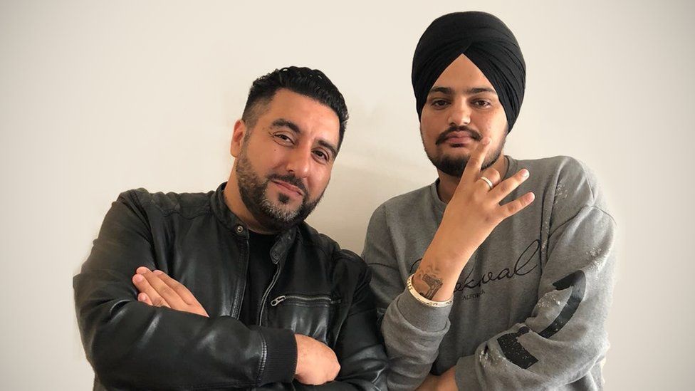 Dipps Bhamrah, wearing a leather jacket, poses with his arms crossed, with a slight smile as he leans in towards Sidhu Moose Walla. Moose Walla's wearing a black turban and grey sweatshirt. He's holding one hand up in a symbol gesture, three fingers are extended but his ring finger is bent.
