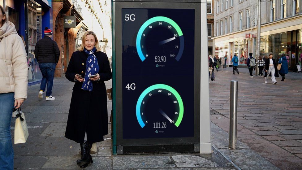Zoe standing beside a pillar with a green screen showing a 4G and 5G speed test in Glasgow city centre