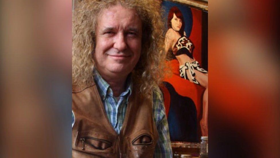 Man with long curly hair in front of a painting of a woman