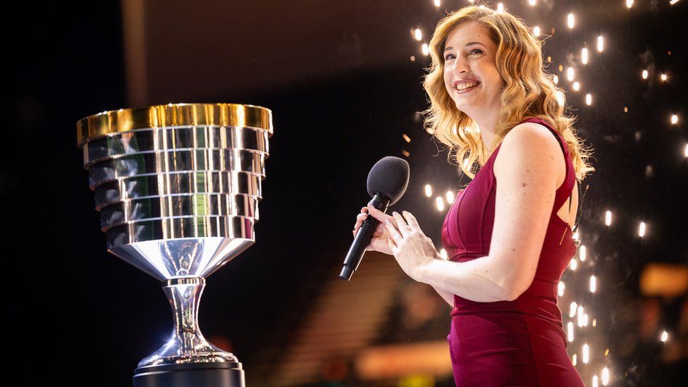 A woman in a red dress holds a microphone, smiling. Behind her, a stream of sparks from a pyrotechnic goes off. She's applauding, looking over in the direction of a large, goblet-shaped trophy. It's mostly silver but has a golden rim which reflects the sparks and stage lights.