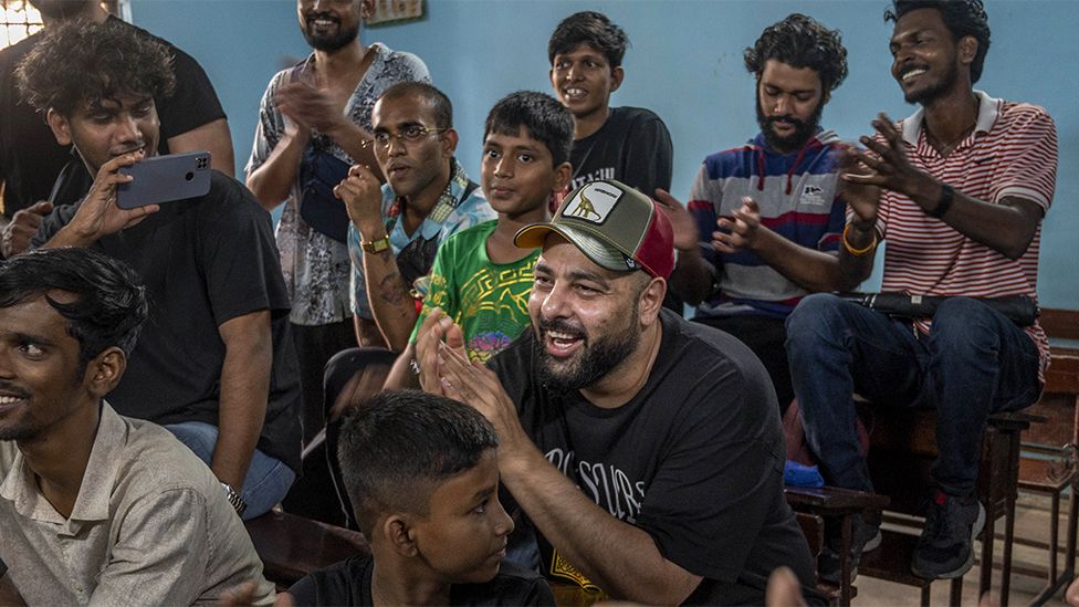 Badshah surrounded by kids as he visits an education project in Mumbai, India. He is wearing a black tshirt with a green and red cap, smiling as his hands are clapping. The kids around him have their phones out recording him, smiling and laughing. The background is a blue wall.