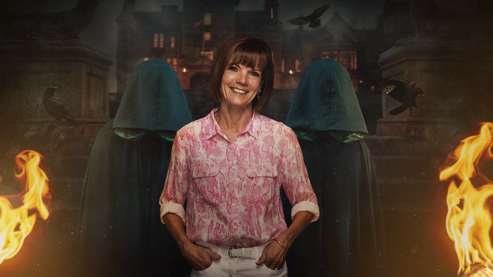 Diane's promotional shot for The Traitors. Diane is a 63-year-old white woman who has a ginger bob. She wears a patterned pink and white blouse over white trousers and has her hands in her pockets while smiling at the camera. She is flanked by two cloaked traitors in front of the Scottish castle where the series is filmed and flames have been added for dramatic effect