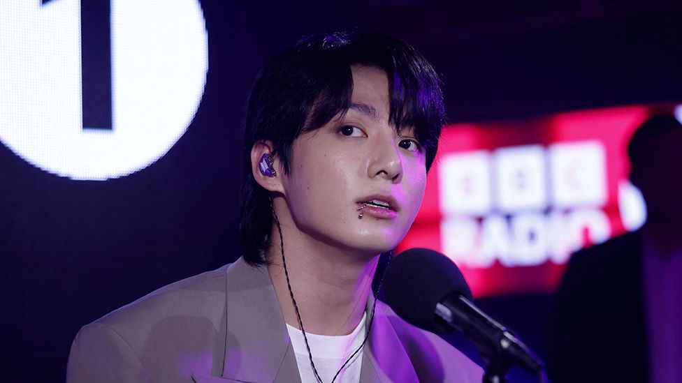 Jung Kook in a beige suit with white t-shirt underneath. He's behind a microphone in a studio, and Radio 1 Live Lounge branding on the wall behind. He looks pensive, as if he's getting ready to perform
