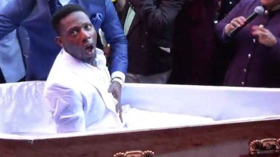Pastor Alph Lukau stands next to the coffin and places his hand on the stomach of the man he is claiming to resurrect.
