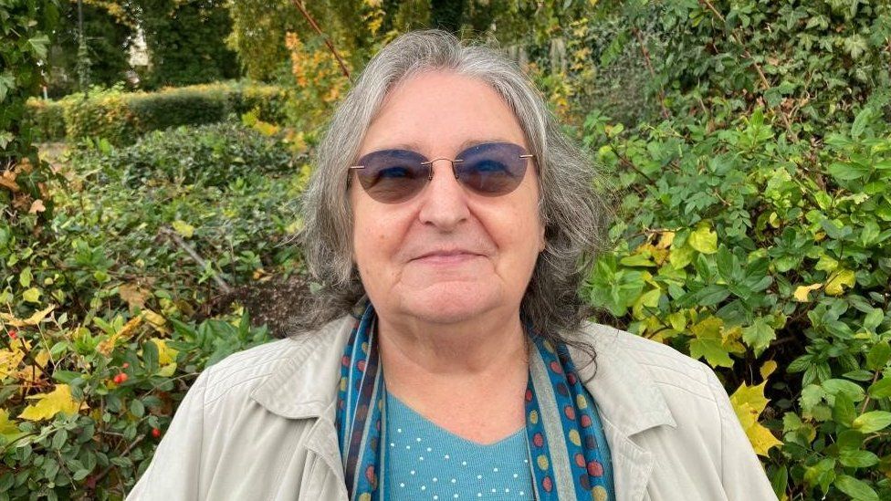 Woman with medium-length grey hair and dark glasses standing in front of a hedge