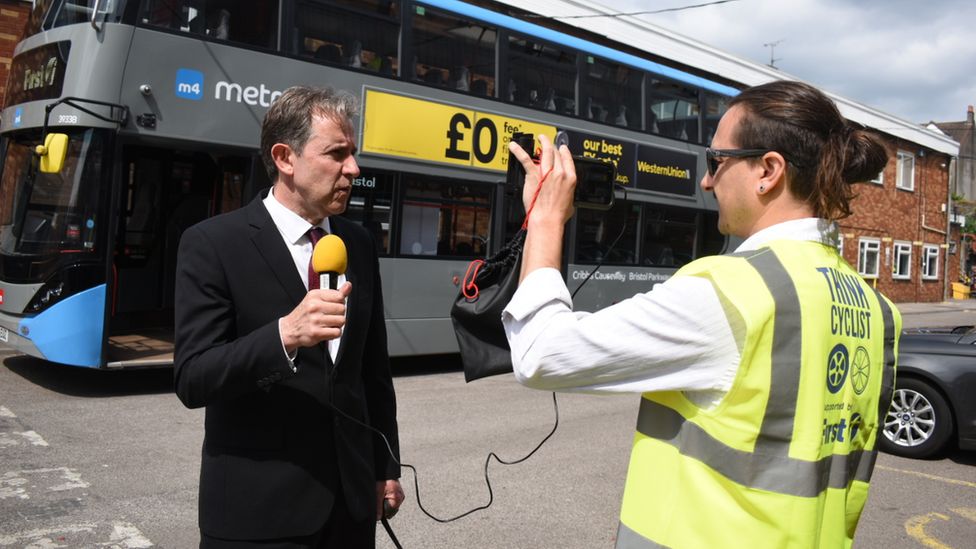 Dan Norris holding a microphone while being filmed in front of a Metrobus