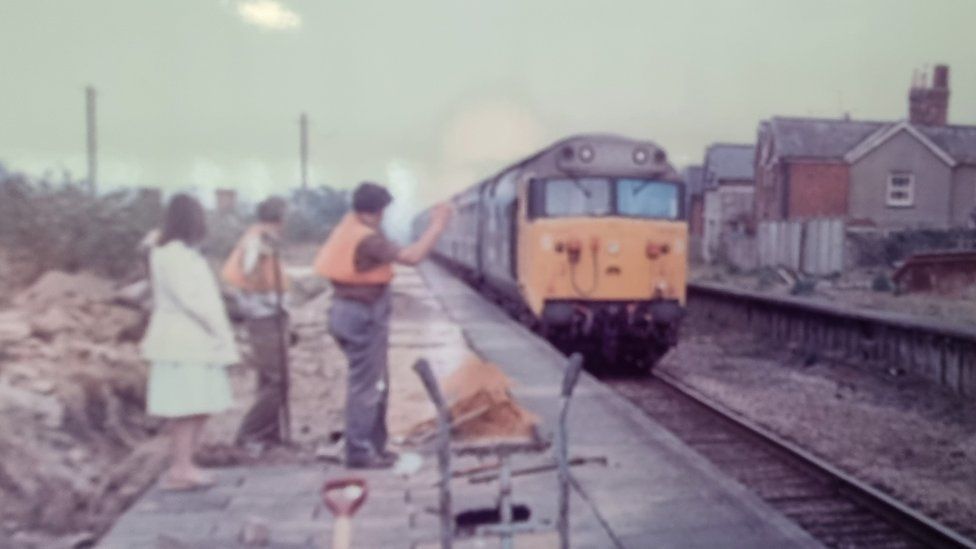An old photograph of Templecombe Station. Three people are standing on the platform waving at a yellow train.