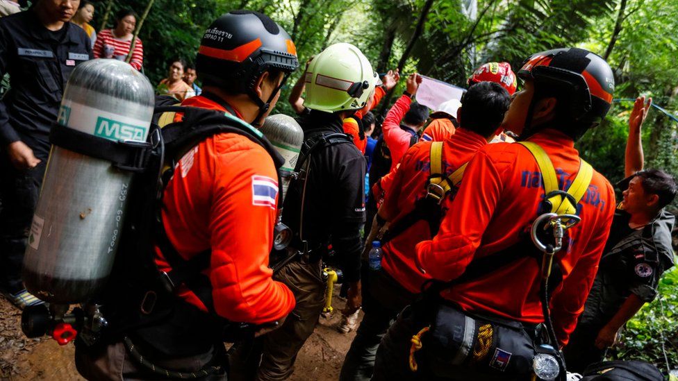 Rescue personnel carrying diving tanks on their backs gather at the cave site