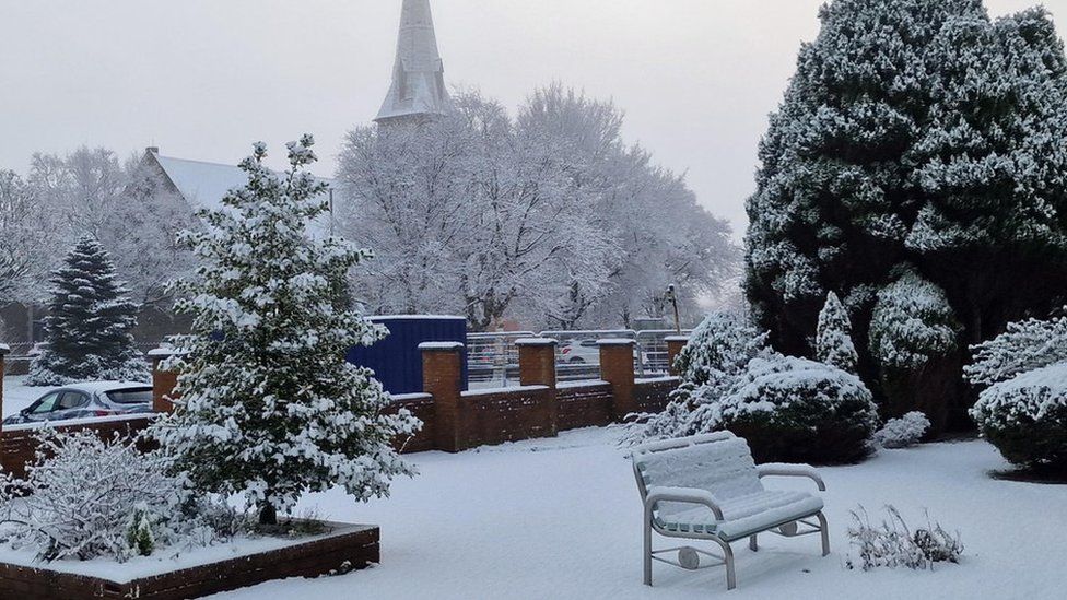 Snow covers a garden and garden furniture with a church spire in the distance.