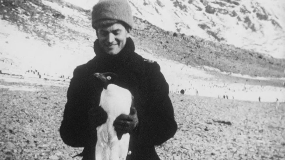 Black and white footage of a man wearing a hat and holding a penguin.