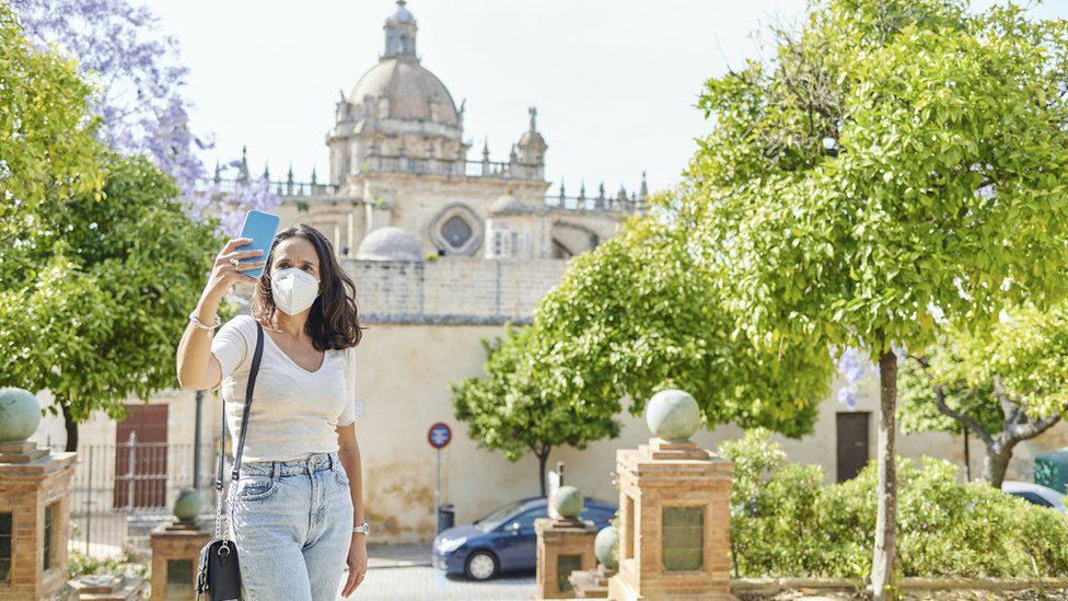 A woman wearing an N95 mask poses for a selfie next to a tourist attraction in Spain