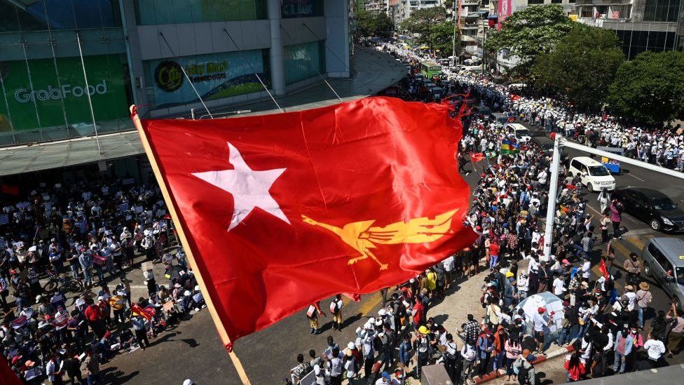The flag of the National League for Democracy party flies over protesters taking part in a demonstration against the February 1 military coup in Yangon