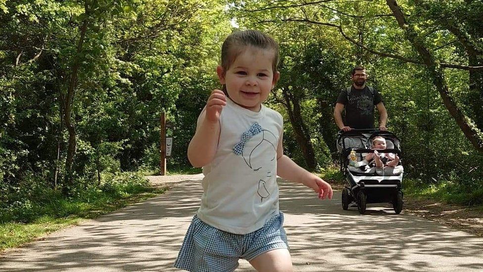 Grace running in park with father and stroller in background