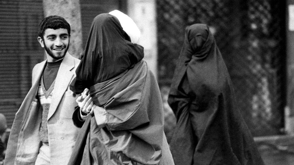 A mullah in a white turban, holding his little daughter in black chador in his arms, talks to a man, while accompanied by his wife, also in black chador with a net mask covering her face, as they head towards Friday prayers at Tehran university, 1 February 1980