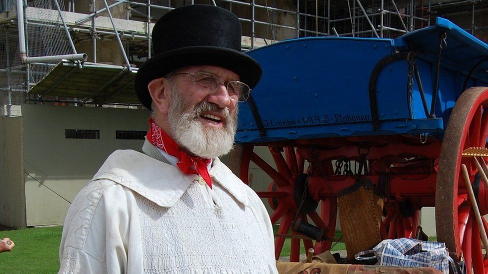 Eric Freeman in a top hat wearing white with a red neckerchief at a history festival. He stands in front of an old blue wagon.
