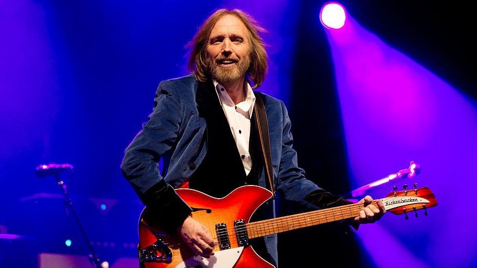 Tom Petty performing at the Isle of Wight Festival in 2012. Tom is in his 60s, still with shoulder length fair hair and a short beard. He wears a white shirt and a blue velvet suit. He smiles as he holds his electric guitar which is orange and red. The staging behind him is lit blue and purple