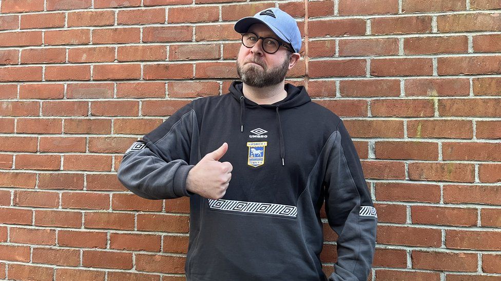 Richard Woodward, wearing an Ipswich Town hoodie and cap, in front of a brick wall