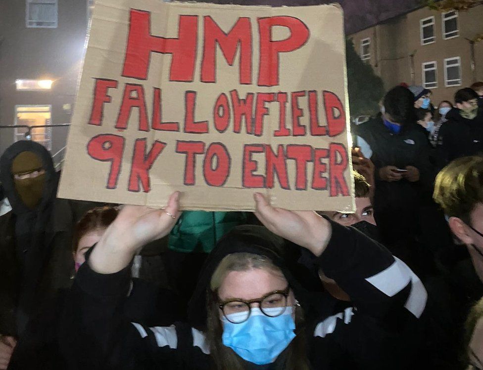 A student holds a sign reading "HMP Fallowfield, 9k to enter"