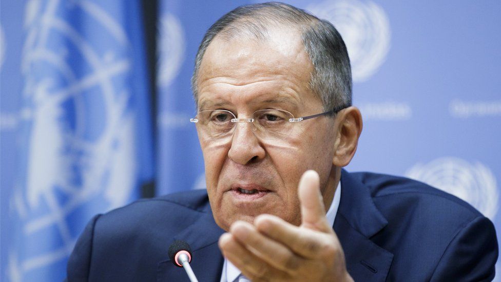 Sergei Lavrov gestures at the camera with a soft-focus UN flag visible in the background
